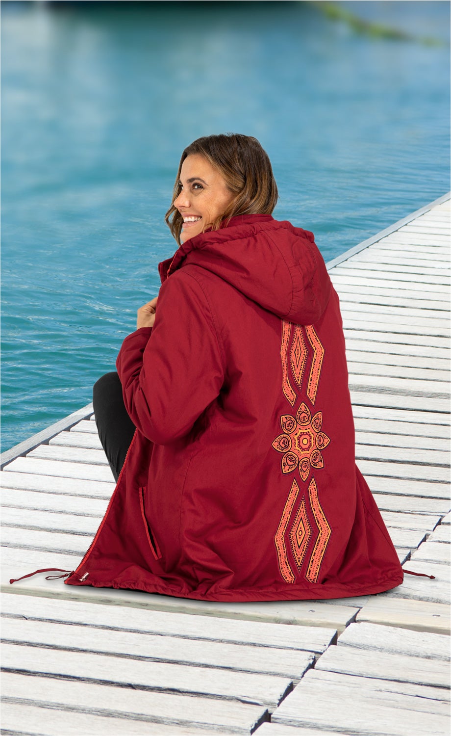 Frau sitzt lächelnd auf Holzsteg am Wasser, trägt rote Baumwoll-Jacke mit Kapuze und orangefarbenem, geometrischem Rückenmuster, schwarze Hose, ruhige Wasserkulisse im Hintergrund
