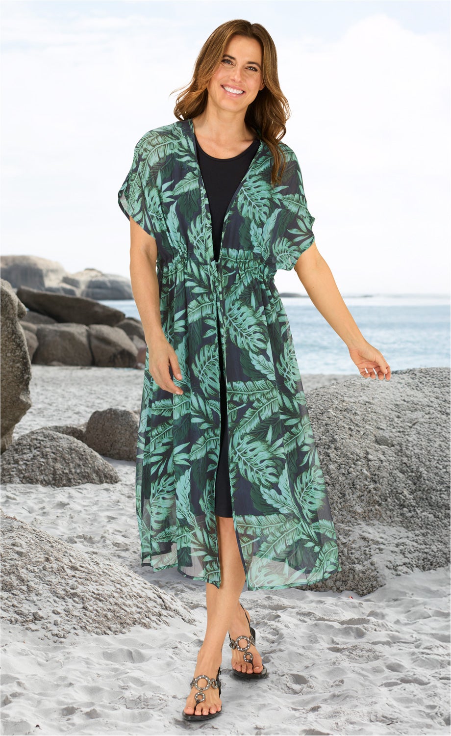 Frau am Strand trägt schwarzen Unterkleid mit darüber offenem, halblangem Viskose-Kaftan mit grünem, tropischem Blattmuster, schwarze Sandalen mit Schmuckringen, lächelnd, braune schulterlange Haare, heller Sand, Felsen und Meer im Hintergrund