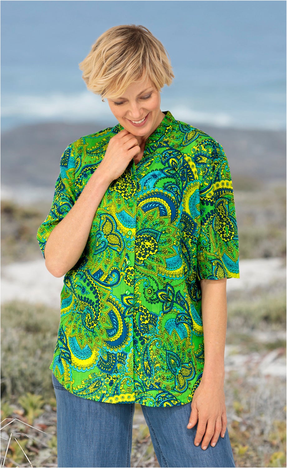 Frau trägt farbenfrohe Viskose-Bluse mit grün-blau-gelbem Paisley-Muster, halblangen Ärmeln und Knopfleiste, kombiniert mit blauer Hose, steht im Freien vor unscharfem Naturhintergrund