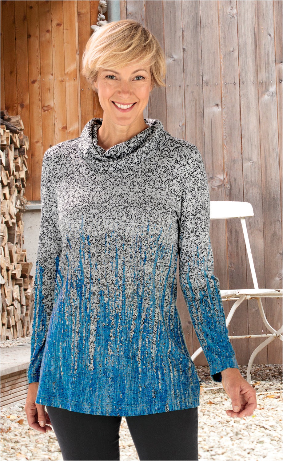 Frau mit kurzem blondem Haar trägt langärmliges Viskose-Mix-Shirt mit grauem Ornamentmuster und blauen Verläufen, steht vor Holzwand mit Holzstapel und weißem Gartenstuhl, lächelnd
