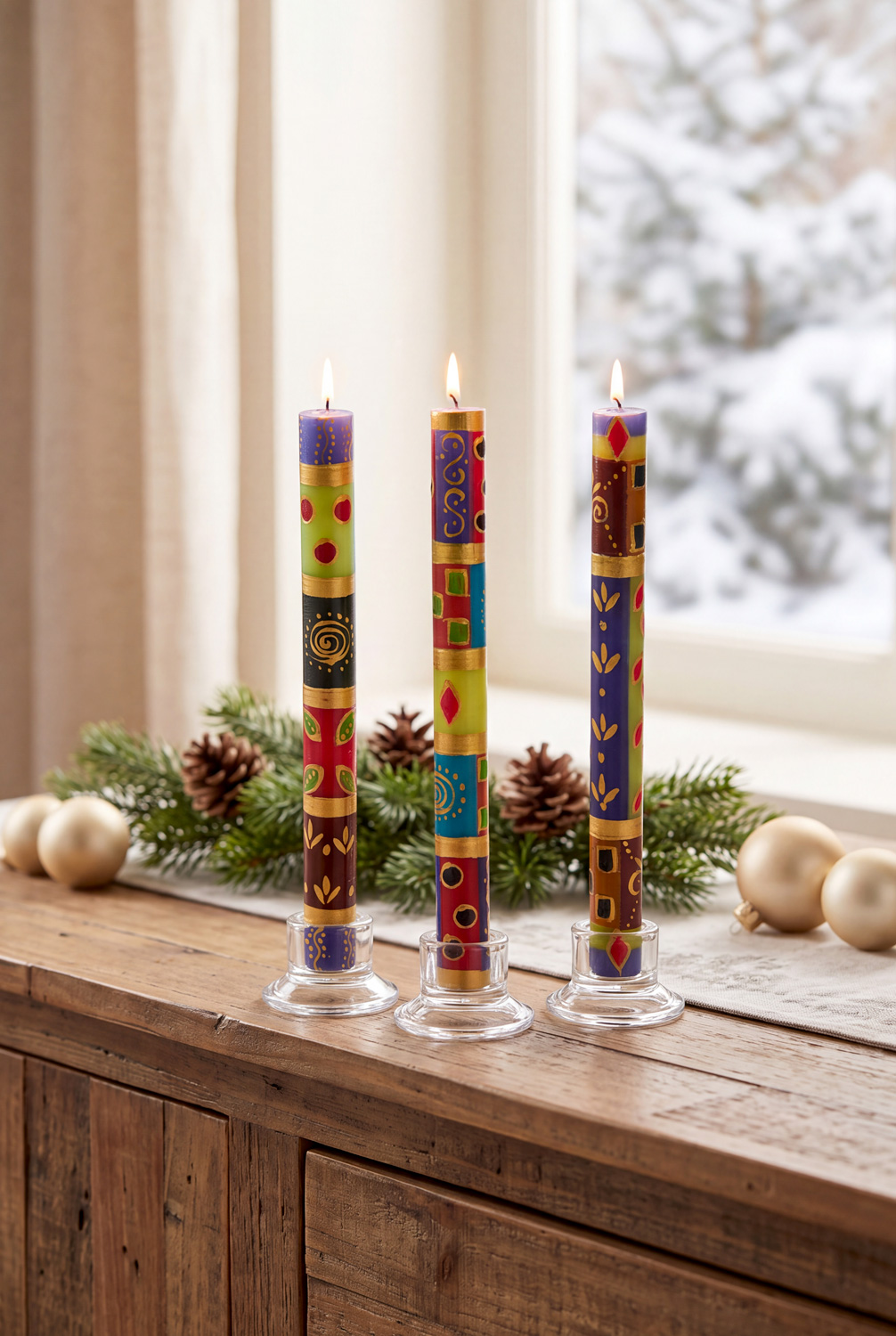 Drei bunte, handsignierte Adventskerzen mit Flamme in transparenten Halterungen auf Holztisch, dahinter Tannenzweige, Zapfen, goldene Weihnachtskugeln, im Hintergrund verschneite Fensterlandschaft und cremefarbener Vorhang