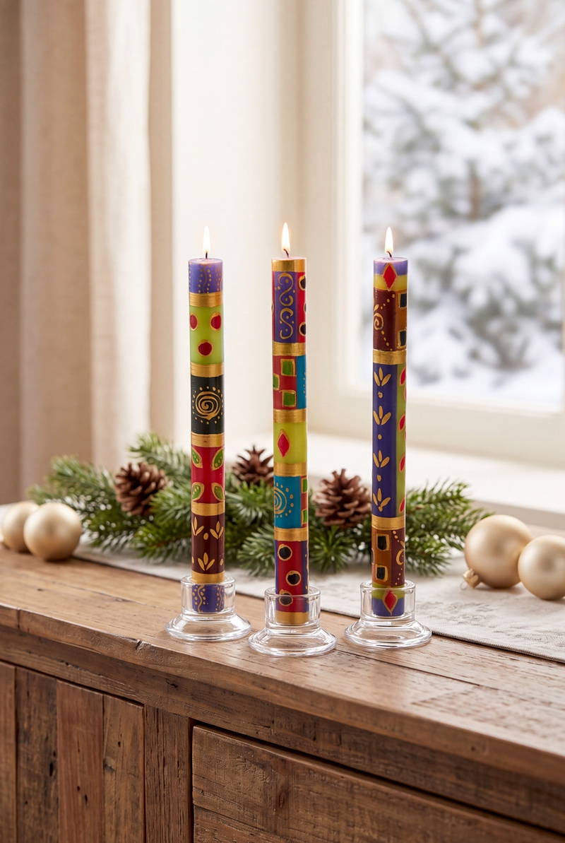 Drei bunte, handsignierte Adventskerzen mit Flamme in transparenten Halterungen auf Holztisch, dahinter Tannenzweige, Zapfen, goldene Weihnachtskugeln, im Hintergrund verschneite Fensterlandschaft und cremefarbener Vorhang