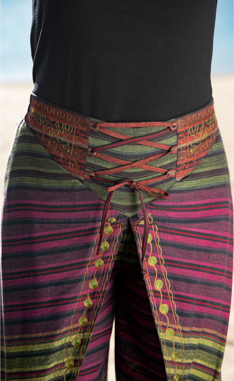 Baumwoll-Hose mit bunten horizontalen Streifen in Grün, Rosa und Schwarz, dekorativen grünen Stickereien an den Beinen, geschnürtem Bund mit roten Bändern und braunen Ziernähten, getragen zu schwarzem Oberteil