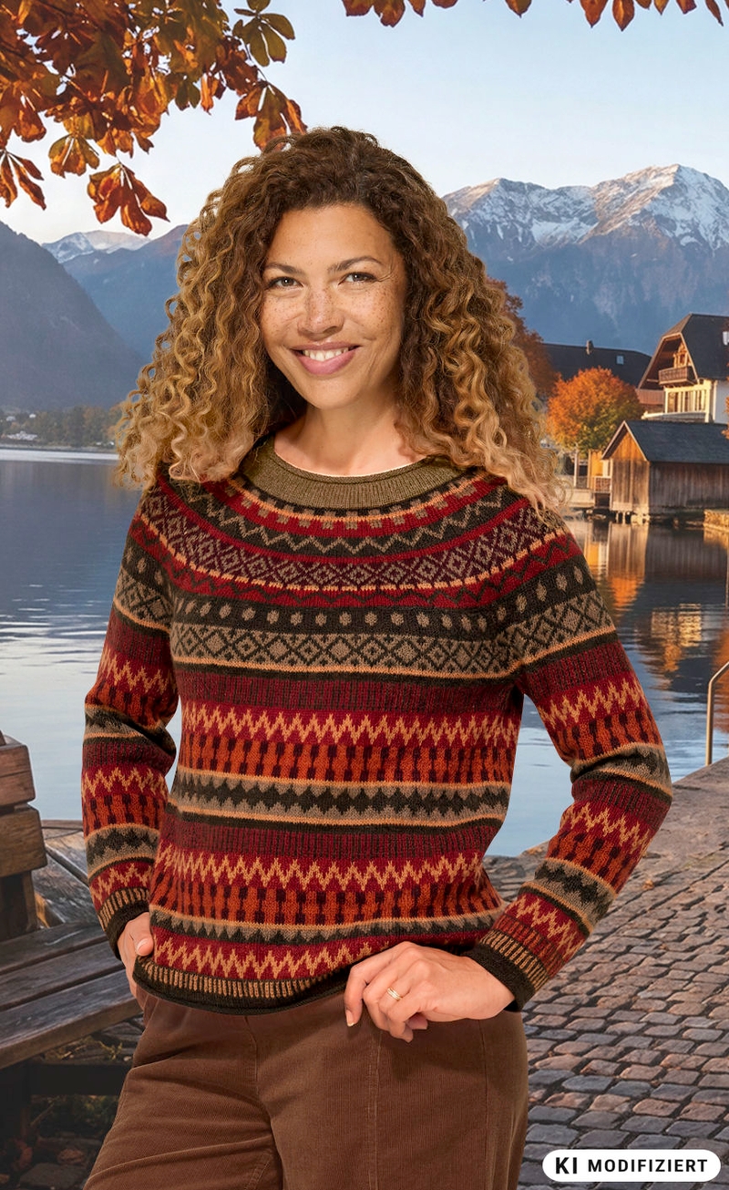 Frau mit lockigem Haar trägt bunt gemusterten Alpaka-Pullover in warmen Herbstfarben, steht an Seeufer mit Steg, im Hintergrund Herbstbäume und Berge mit Schnee, sonniger Himmel, herbstliche Atmosphäre