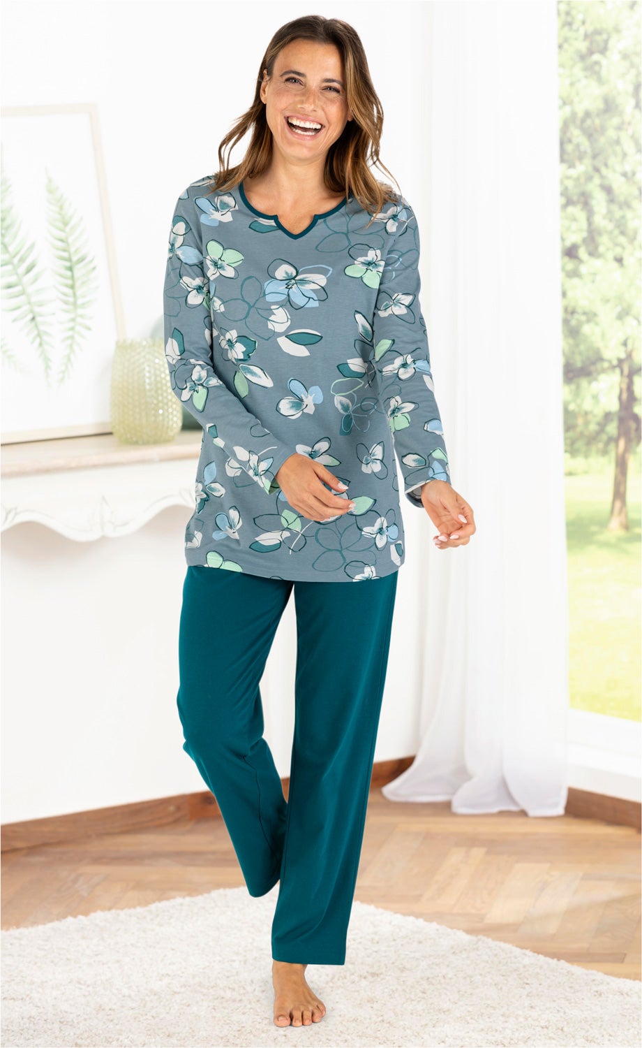 Frau trägt Baumwoll-Pyjama Set mit blau-grünem, floralem Oberteil und einfarbiger, dunkelgrüner Hose, steht barfuß auf hellem Teppich in hellem Raum mit Fenster und Pflanzenbild im Hintergrund, lächelnd und bewegt sich entspannt