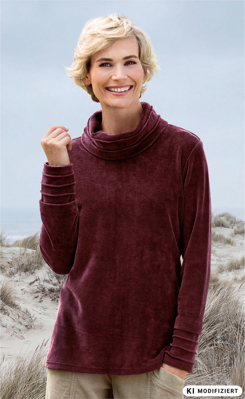 lächelnde Frau mit kurzen blonden Haaren trägt bordeauxfarbenen Rollkragenpullover aus Samt und helle Hose, steht vor Dünenlandschaft am Strand bei bewölktem Himmel