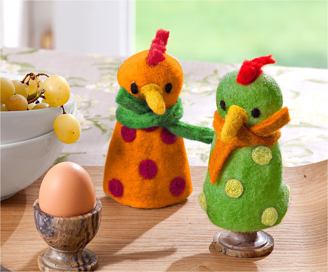 Zwei bunte Eierwärmer aus Filz in Form von Vögeln, einer orange mit roten Punkten und grünem Schal, der andere grün mit gelben Punkten und orangefarbenem Schal, jeweils auf einem Marmor-Eierbecher stehend, daneben ein braunes Ei und eine weiße Schale mit