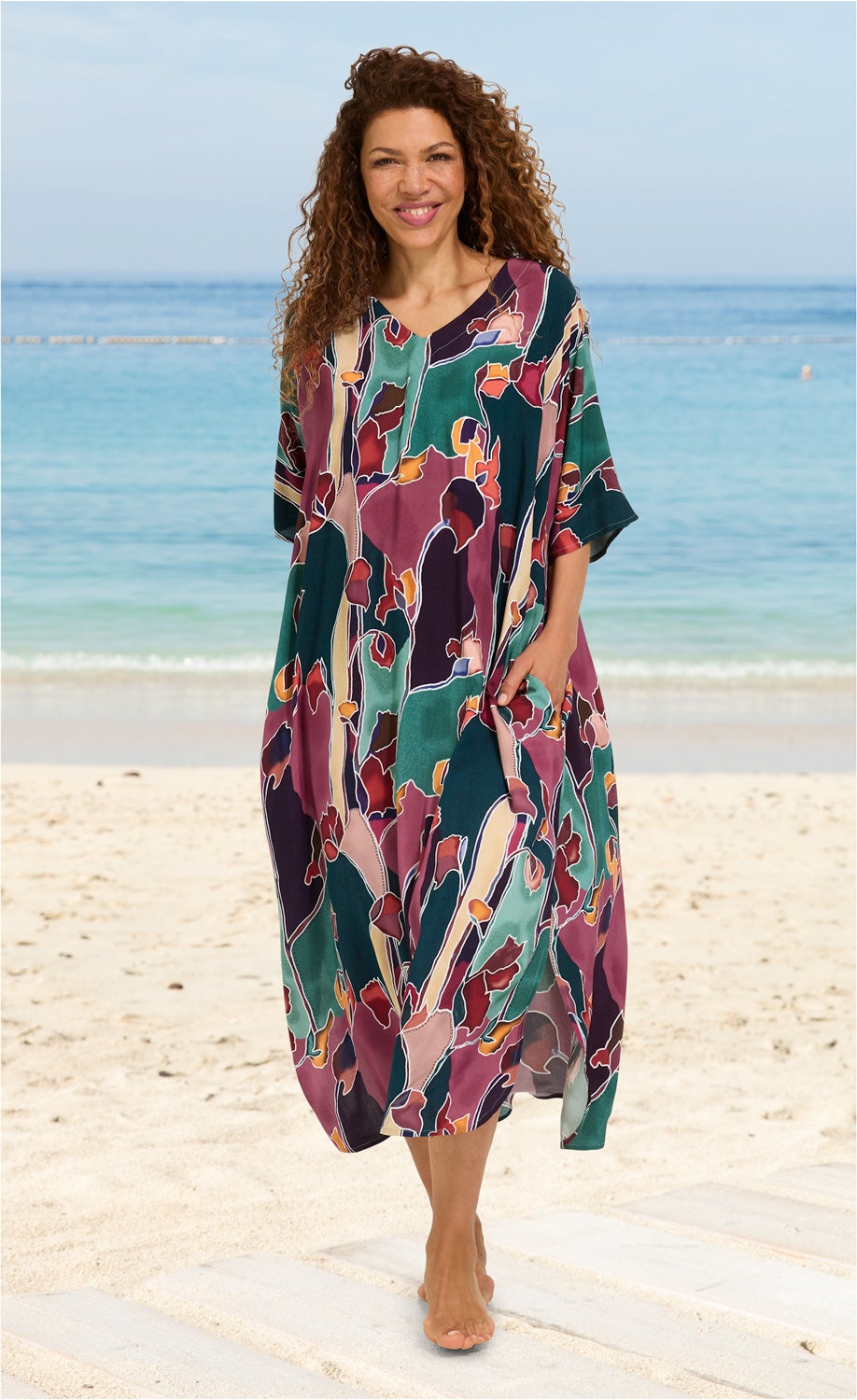 Frau am Strand trägt bunten Viskose-Kaftan mit abstraktem Muster in Grün, Lila, Rosa und Orange, halblange Ärmel, V-Ausschnitt, barfuß, lockige Haare, lächelnd, Hintergrund Meer und Sandstrand