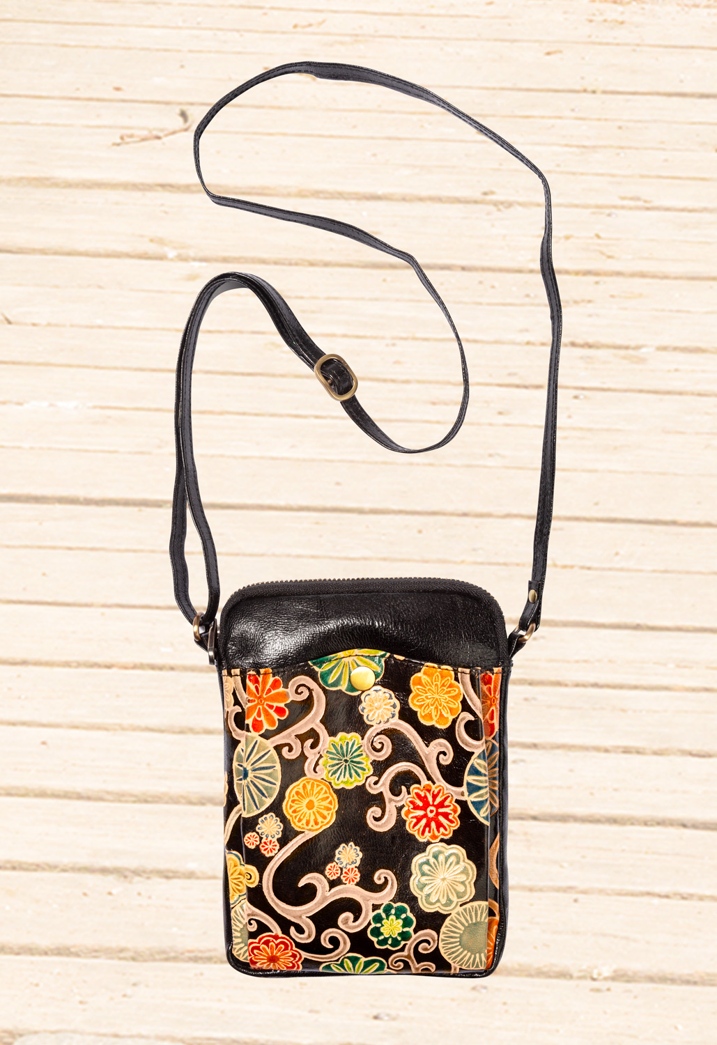 Kleine schwarze Leder-Tasche mit verstellbarem Schulterriemen, frontaler Außentasche mit farbenfrohem floralen Muster in Rot, Gelb, Grün und Beige, goldfarbener Druckknopfverschluss, strukturierter Hintergrund aus hellem Holz