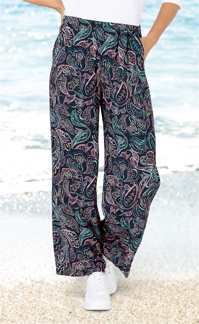 Helle Viskose-Hose mit weitem Bein und buntem Paisley-Muster in Blau, Rosa und Grün, Model trägt weiße Sneakers, Im Hintergrund Strand und Meer bei sonnigem Wetter