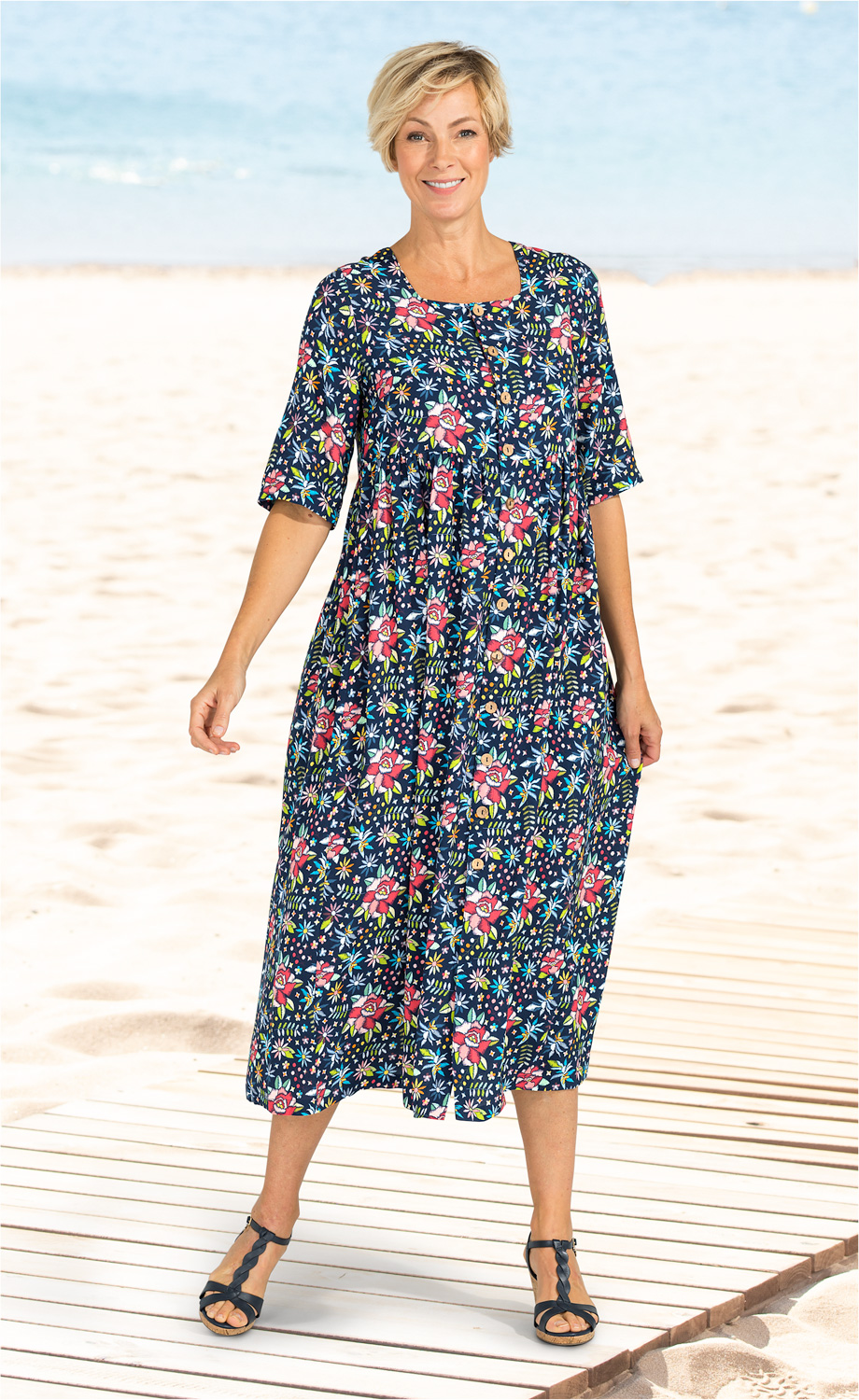 Frau am Strand trägt knielanges, blaues Viskose-Kleid mit buntem Blumenmuster, halblangen Ärmeln und Knopfleiste, dazu schwarze Sandalen mit Riemchen, lächelnd, stehender Pose auf Holzsteg vor Meer und Sandstrand
