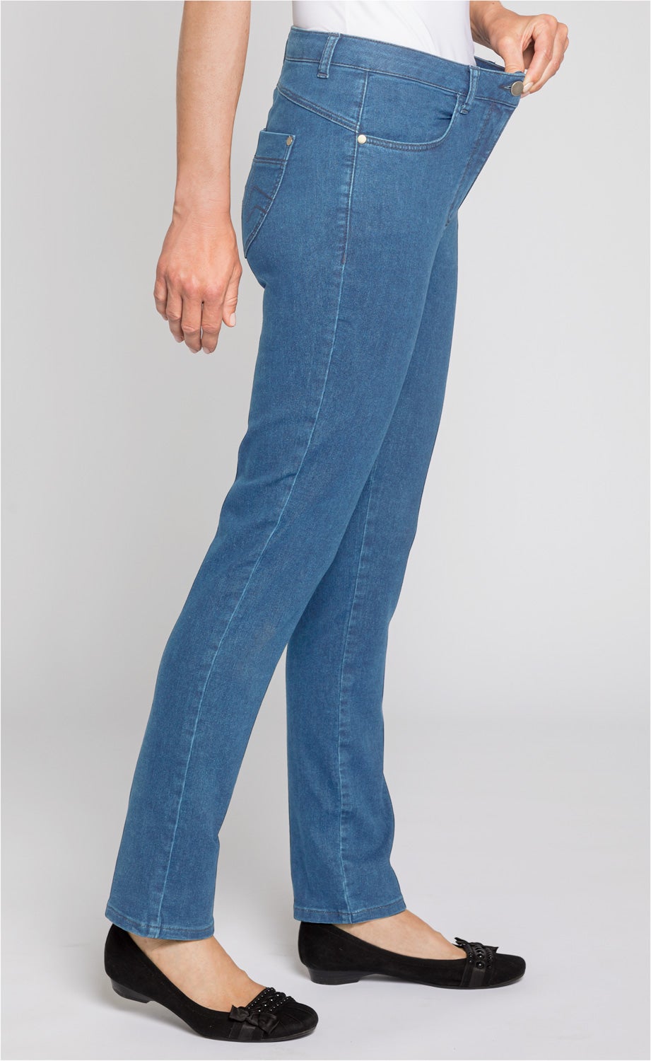 Seitenansicht einer Person, die eine blaue, eng anliegende Baumwoll-Mix-Hose trägt, mit Hand, die den Bund etwas nach außen zieht, kombiniert mit schwarzen Ballerinas mit Schleifen- und Nietenverzierung.