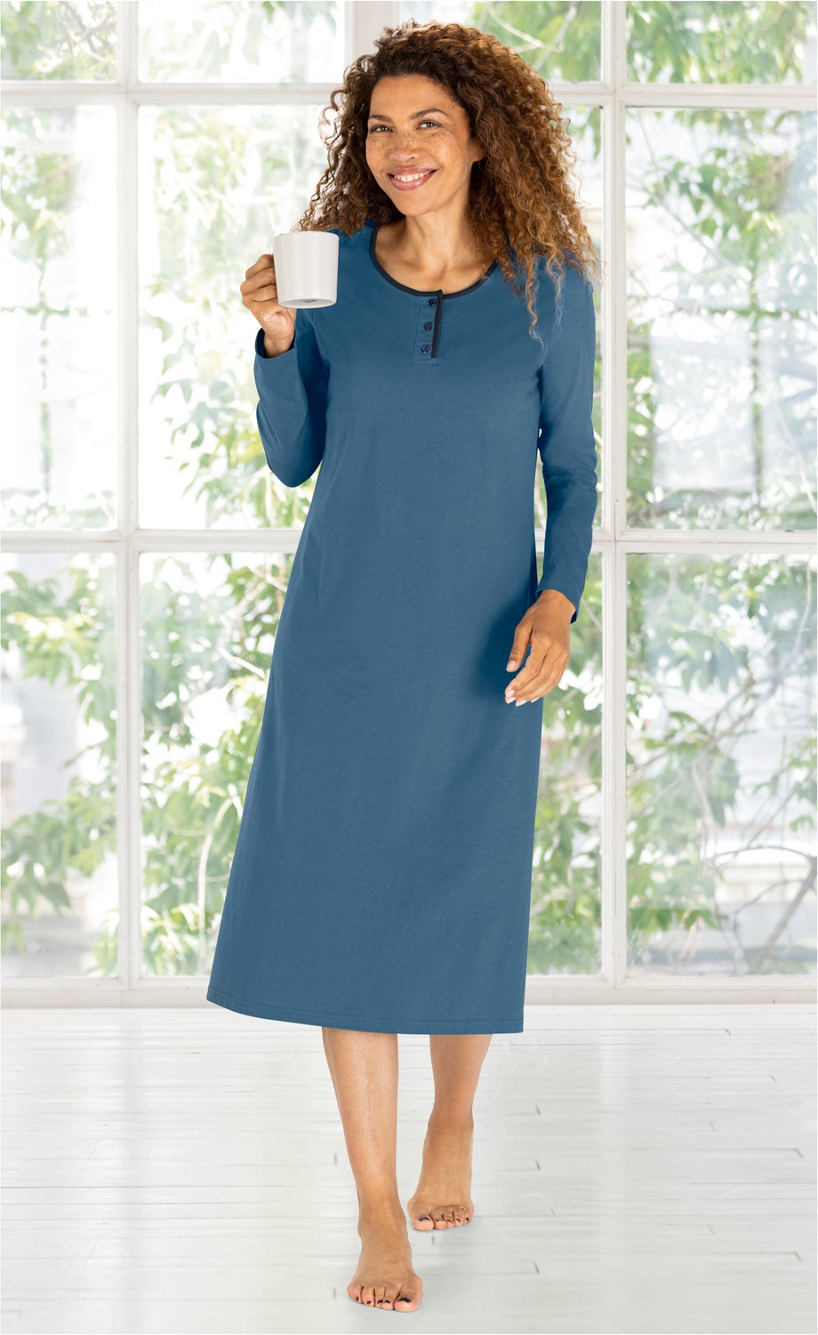 Frau in blauem Baumwoll-Nachthemd mit langem Arm, lächelnd, hält weiße Tasse, steht barfuß in hellem Raum mit großen Fenstern und grünem Pflanzenhintergrund, entspannte Morgenstimmung.