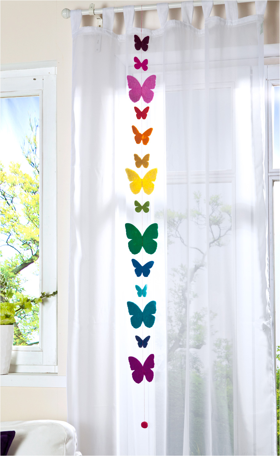 Girlande aus Filz in Form von Schmetterlingen in verschiedenen Farben von Lila, Pink, Rot, Orange, Gelb, Grün bis Blau, aufgehängt vor weißem Vorhang vor Fenster mit Tageslicht und Pflanze unten links.
