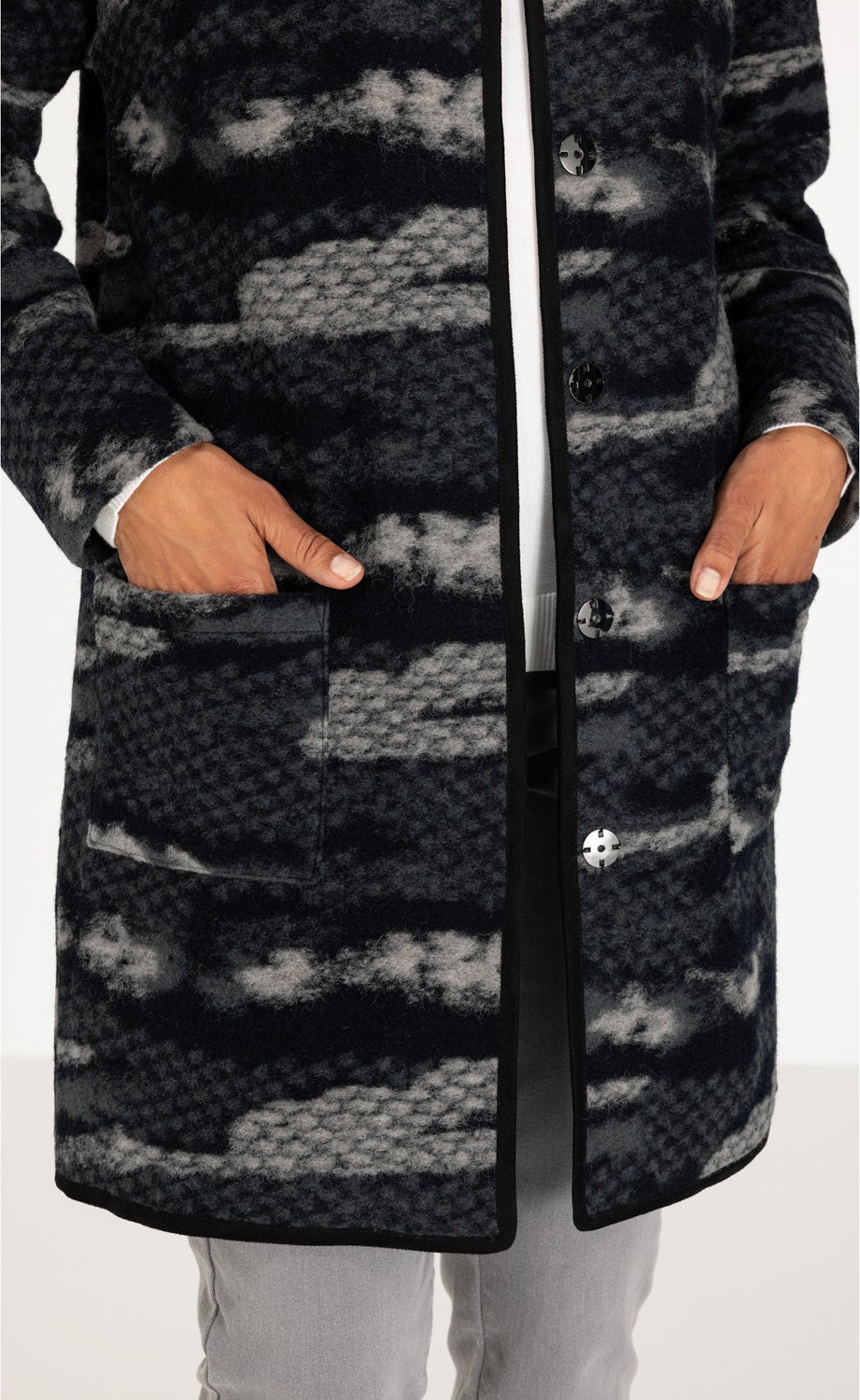 Woll-Mix-Mantel mit grauem und schwarzem Muster, schwarze Knöpfe, zwei aufgesetzte Taschen, schwarze Kantenverarbeitung, getragen mit hellgrauer Hose und weißem Pullover sichtbar an Ärmeln und unter Mantelöffnung