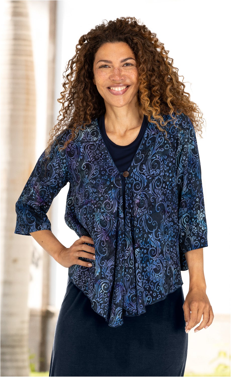 Frau mit lockigem Haar trägt eine blau-schwarze Viskose-Jacke mit floralem Muster und 3/4-Ärmeln, darüber schwarzes Oberteil und dunkler Rock, steht mit Hand in Hüfte, lächelt, heller unscharfer Hintergrund.