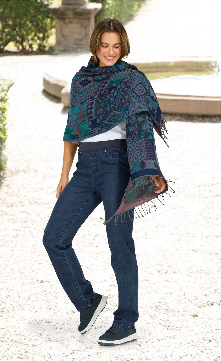 Frau trägt blau-grünen Jacquard-Schal mit geometrischem Muster und Fransen, kombiniert mit weißem Oberteil, blauen Jeans und schwarzen Sneakern, steht lächelnd im hellen Garten mit Springbrunnen im Hintergrund