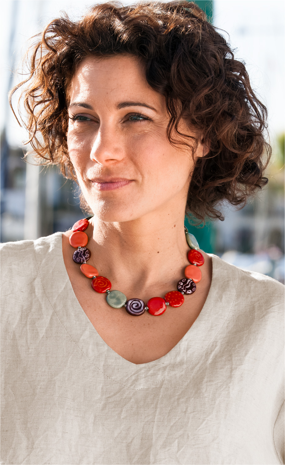 Frau mit kurzen lockigen braunen Haaren trägt eine bunte Keramik-Kette mit flachen, ovalen Perlen in Rot, Orange, Lila und Grün, Musterdetails, dazu schlichtes, helles Leinenoberteil, natürlicher Tageslicht-Hintergrund.