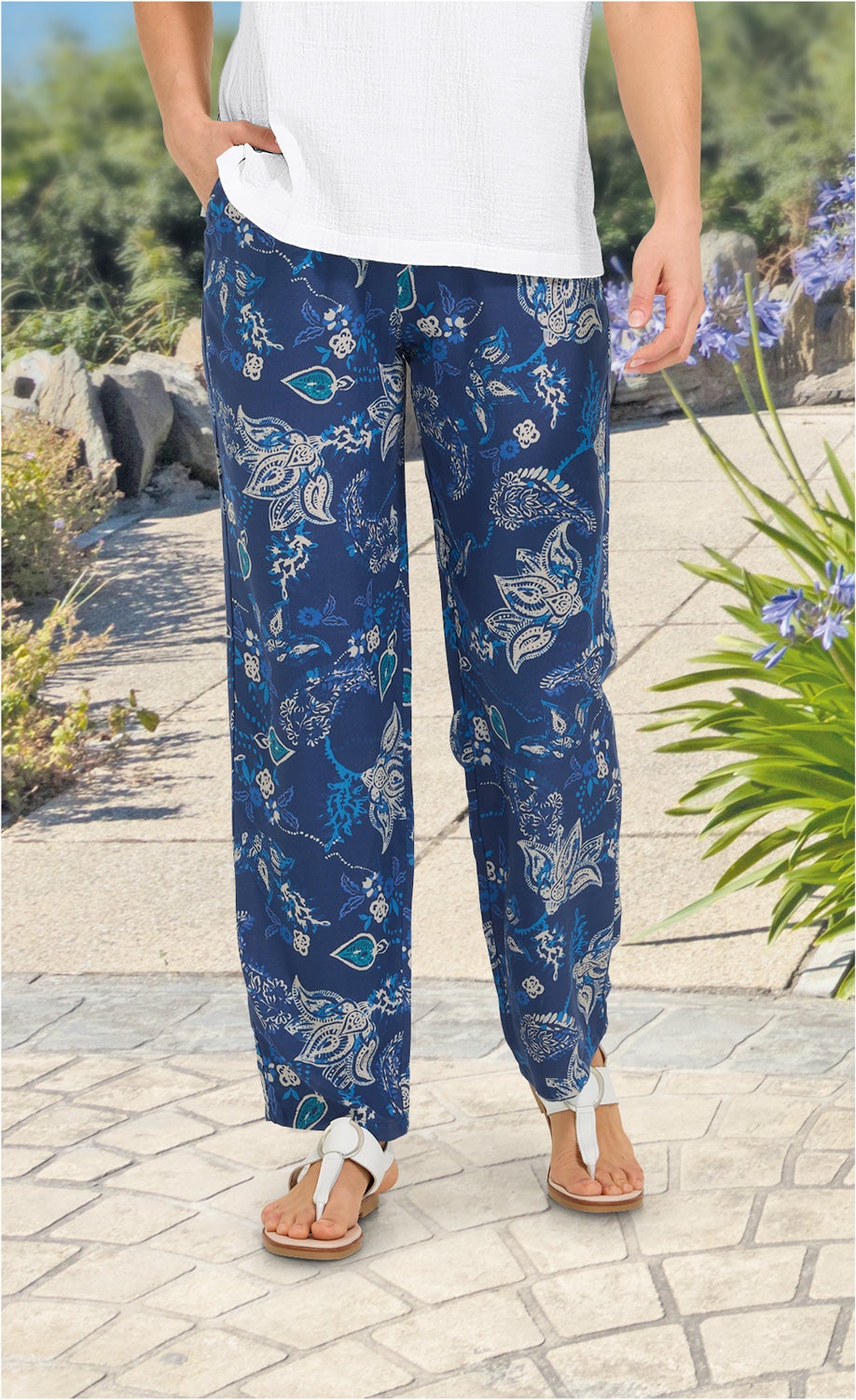 Beinaufnahme einer Person mit weißen Sandalen und einer dunkelblauen Viskose-Hose mit weiß-blauem Blumenmuster, kombiniert mit einem weißen Oberteil, stehend auf gepflastertem Weg mit Pflanzen im Hintergrund