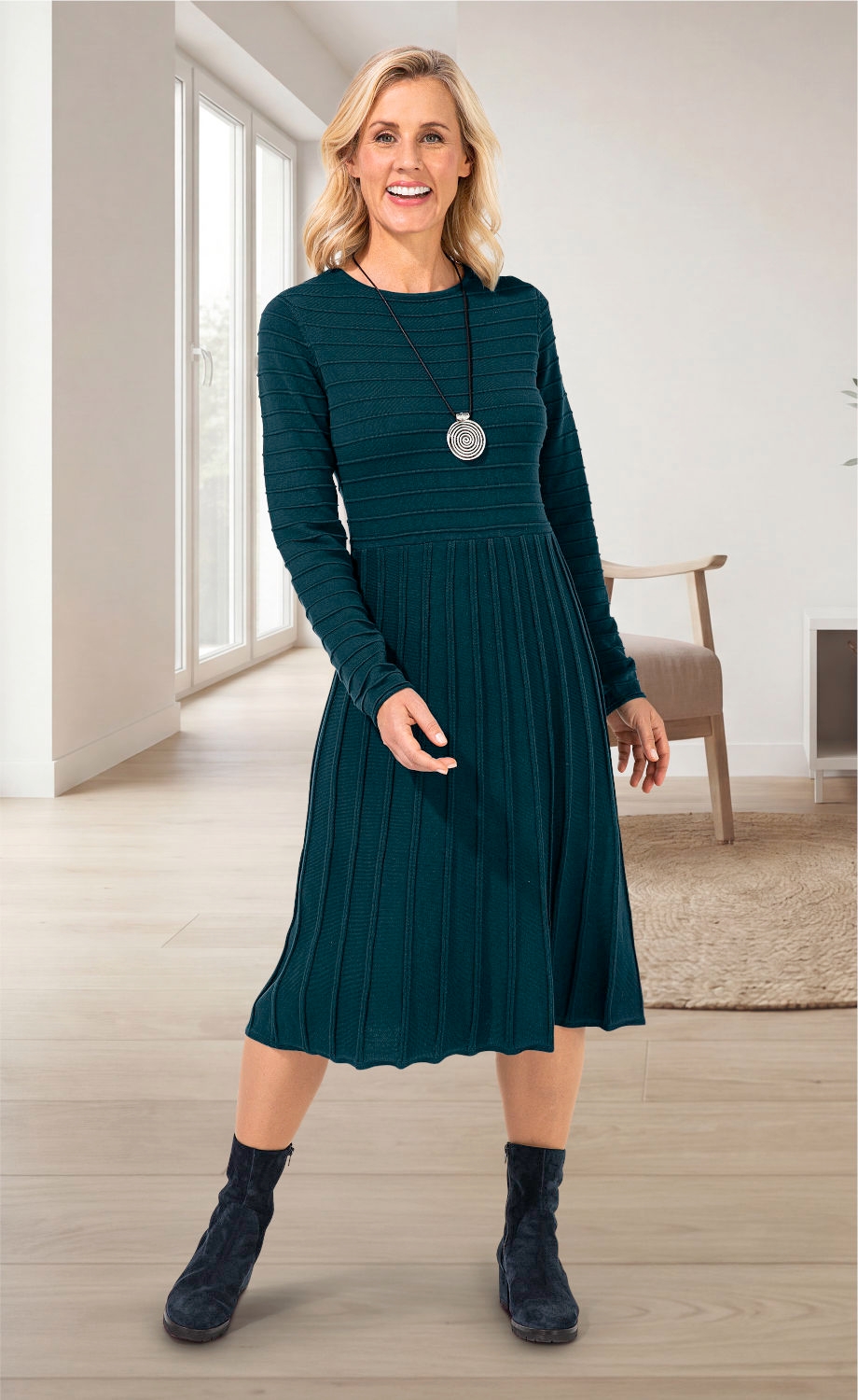 Frau trägt ärmelloses, dunkelgrünes Baumwoll-Kleid mit feinen Streifen und Faltenrock, schwarze Stiefeletten, silberne Spiral-Halskette, steht in hellem, modernem Wohnzimmer mit großer Fensterfront und Holzboden