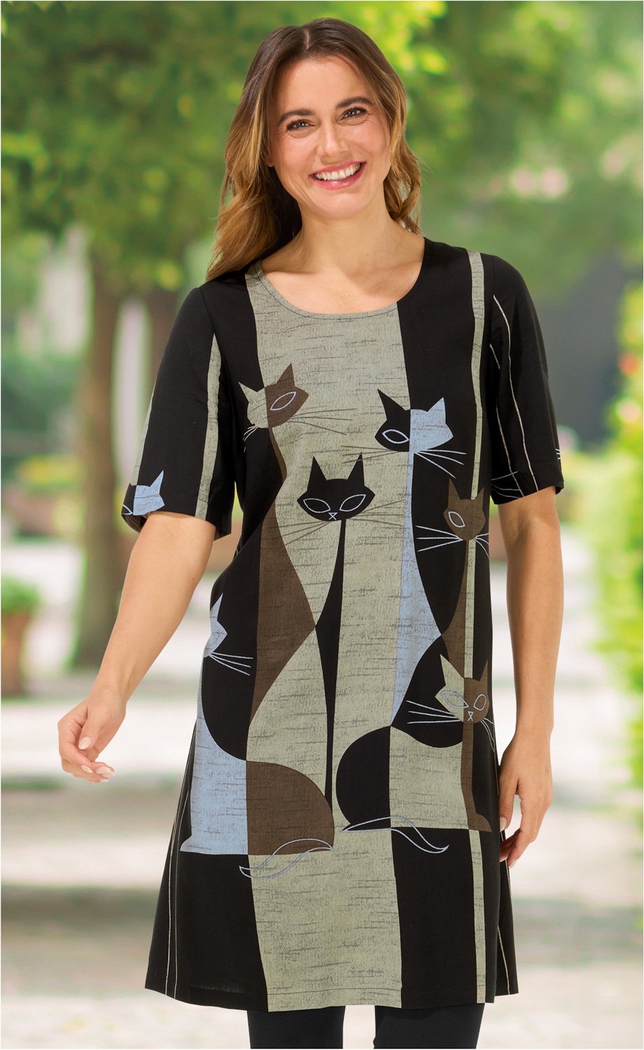 Frau trägt schwarzes Viskose-Kleid mit halblangen Ärmeln, Retro-Katzenmotiv in Braun, Blau und Schwarz, grüner Hintergrund unscharf, lächelnd, mittellange braune Haare