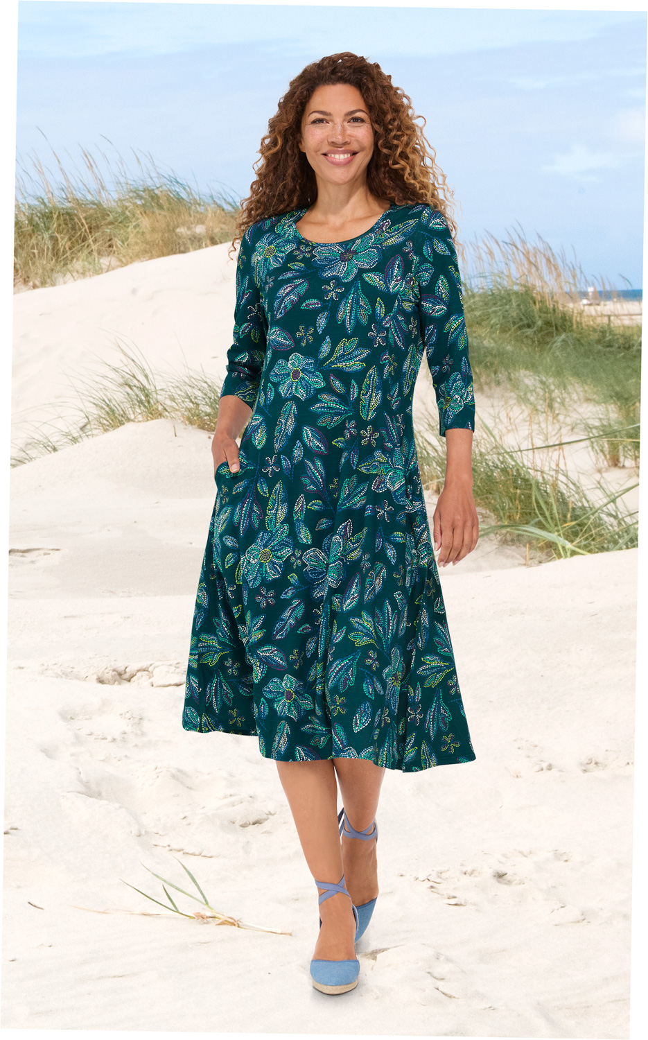 Frau mit lockigen Haaren trägt ein knieumspielendes Viskose-Kleid mit 3/4 Ärmeln und floralen, bunten Mustern, geht barfuß am Strand mit Sanddünen und Gras im Hintergrund, lächelnd, seitliche Taschen vorhanden