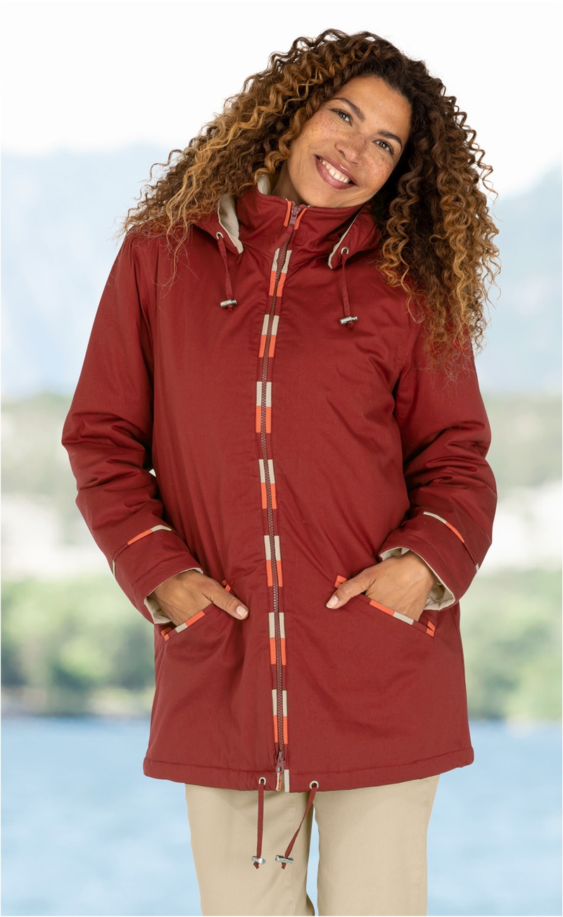 Frau mit lockigen Haaren, lächelt, trägt rote Jacke mit Kapuze und beige Hose, Hände in Taschen, unscharfer Hintergrund mit Wasser und grüner Landschaft.