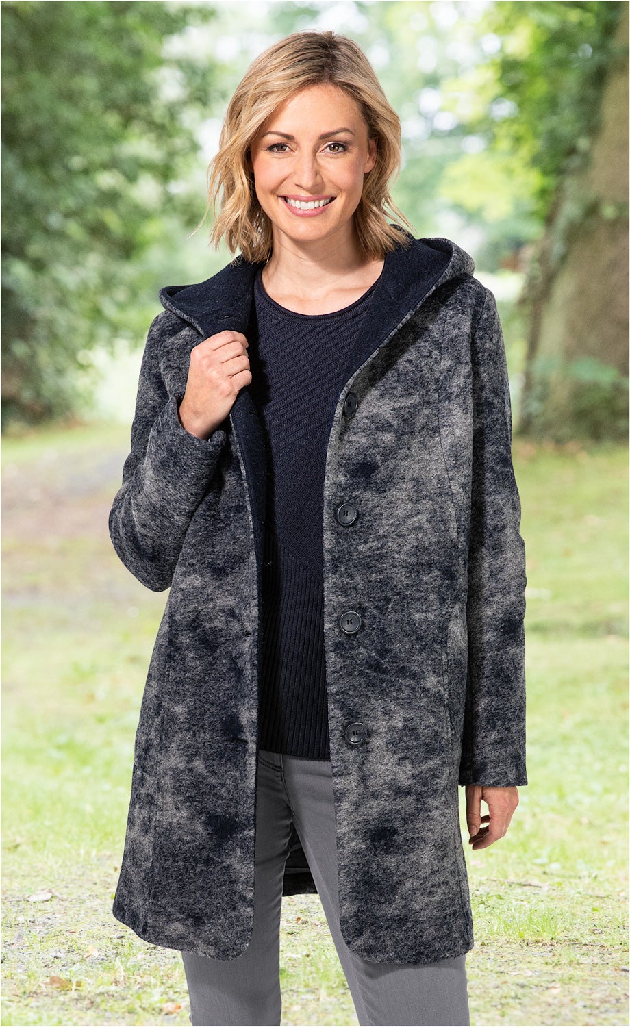 Frau trägt graue Wollmix-Jacke mit Kapuze und Knöpfen, kombiniert mit schwarzem Pullover und grauer Hose, stehend in natürlicher, grüner Parkumgebung, lächelnd