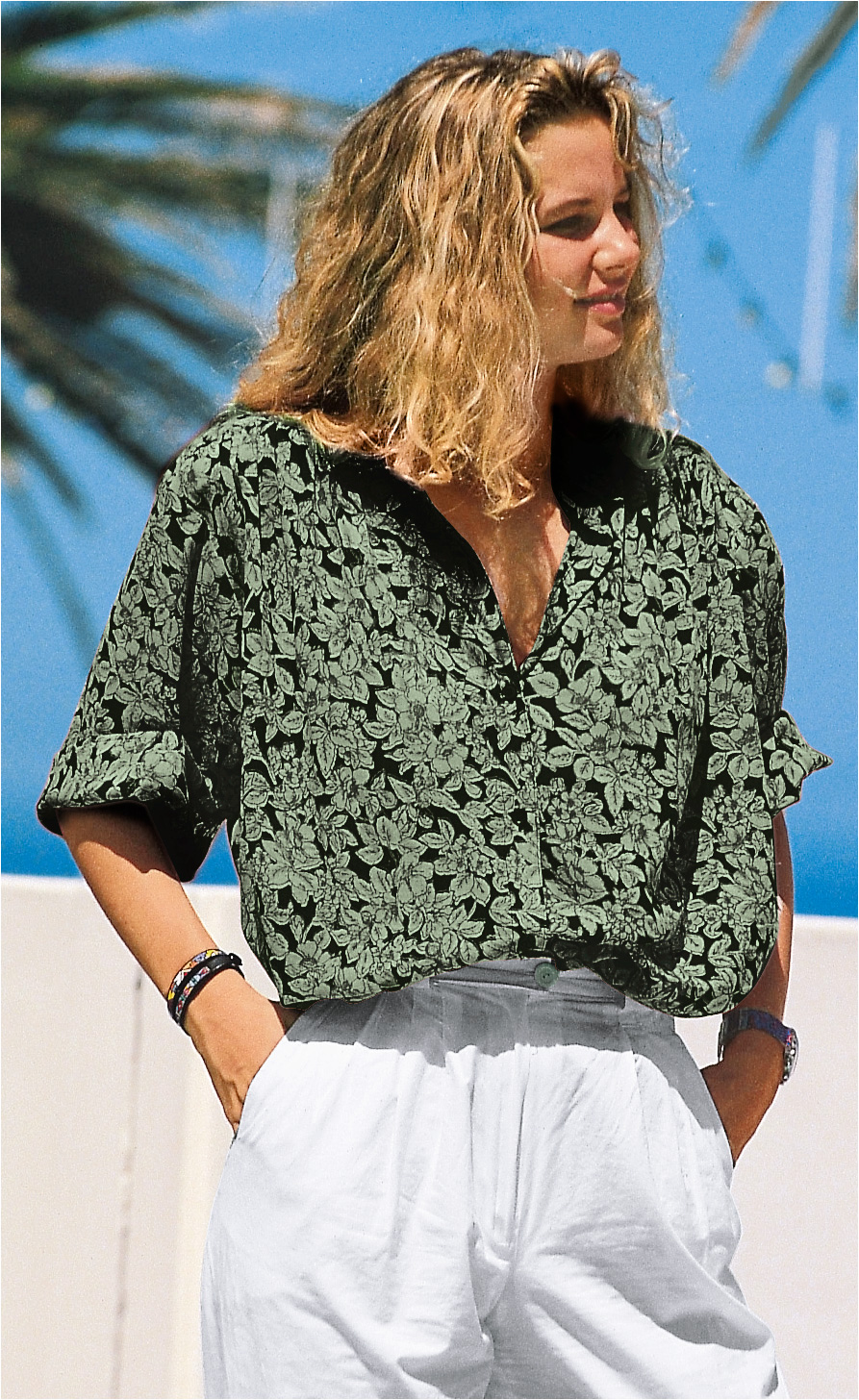 Frau mit lockigem blonden Haar trägt grüne, floral gemusterte Viskose-Bluse mit halblangen Ärmeln und weißen, weiten Hosen vor blauem Himmel und Palmen im Hintergrund