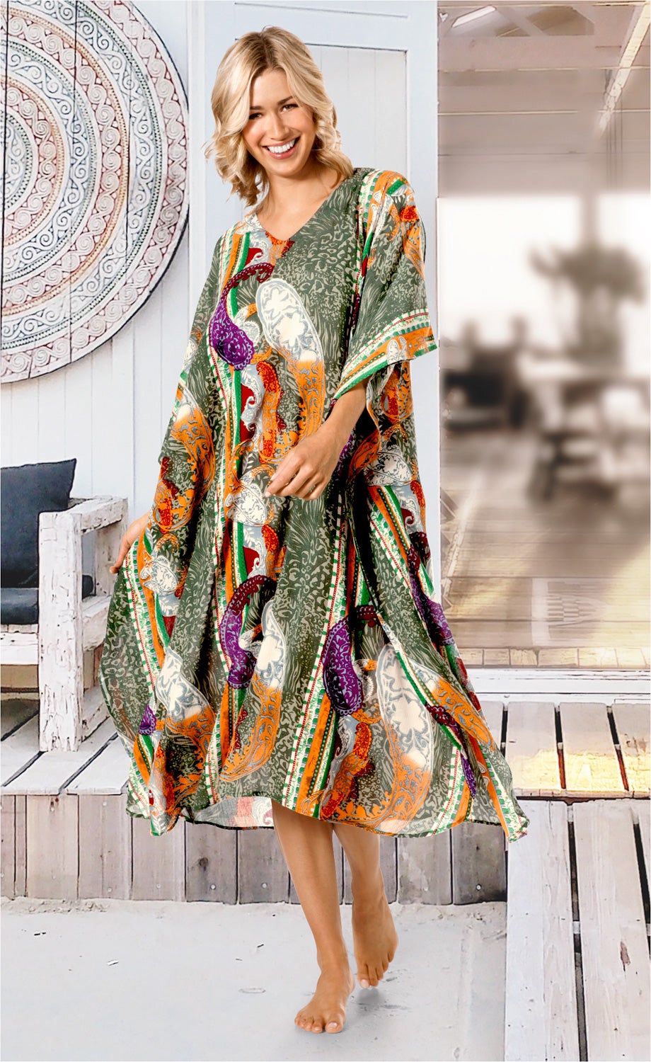 Frau trägt bunten Viskose-Kaftan mit Paisley-Muster in Grün, Orange, Lila und Weiß, lächelt, barfuß, stehend auf Holzterrasse mit Holzmöbeln und dekorativer runder Wandplatte im Hintergrund