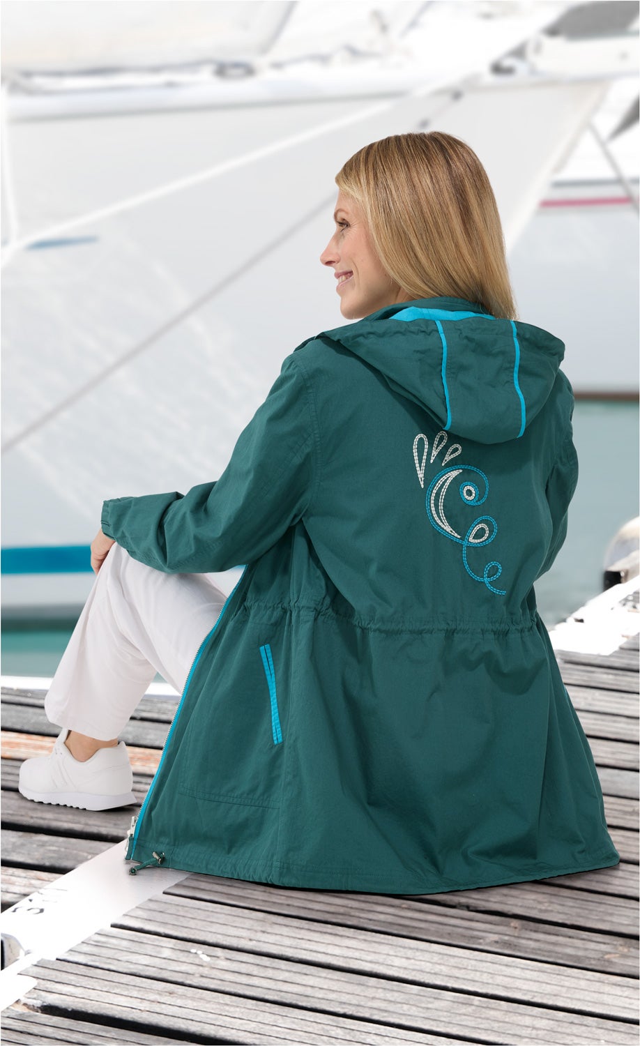Frau sitzt entspannt auf Steg, trägt grüne Baumwoll-Jacke mit Kapuze und blauer Stickerei auf Rücken, weiße Hose und weiße Schuhe, Hintergrund zeigt Wasser und Segelboot teilweise sichtbar