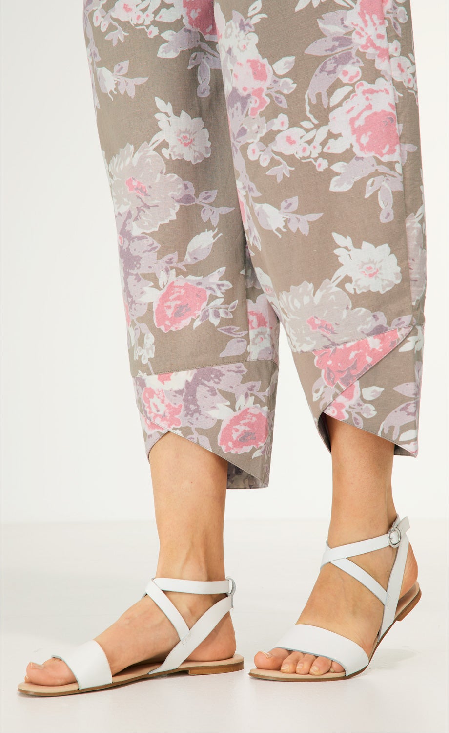 Beine mit Baumwoll-Mix-Hose in Beige mit rosa-weißem Blumenmuster und asymmetrischem Saum, kombiniert mit weißen, flachen Sandalen mit Riemen und Schnalle.
