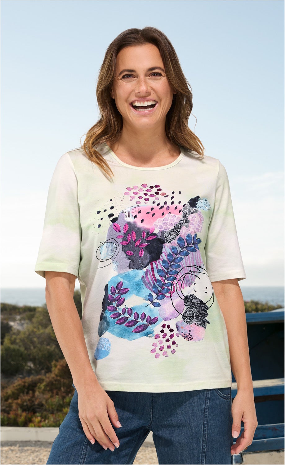 Frau trägt cremefarbenes Baumwoll-Shirt mit buntem abstraktem Blatt- und Muster-Print in Blau-, Lila- und Pinktönen, kombiniert mit blauer Jeans, lächelt, steht vor hellem Himmel und natürlicher Landschaft im Hintergrund
