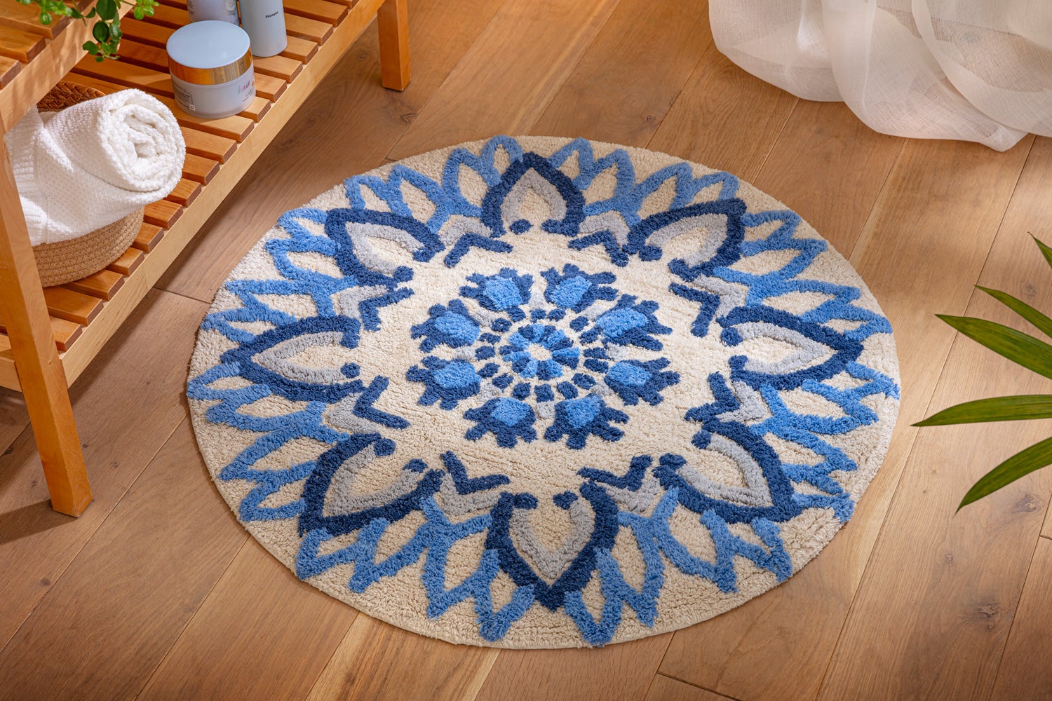 Runde Baumwoll-Badematte mit 75 cm Durchmesser, cremefarben mit blauem und dunkelblauem floralen Ornamentmuster, auf hellem Holzboden platziert, neben einem Holzregal mit weißen Handtüchern und Kosmetikartikeln.