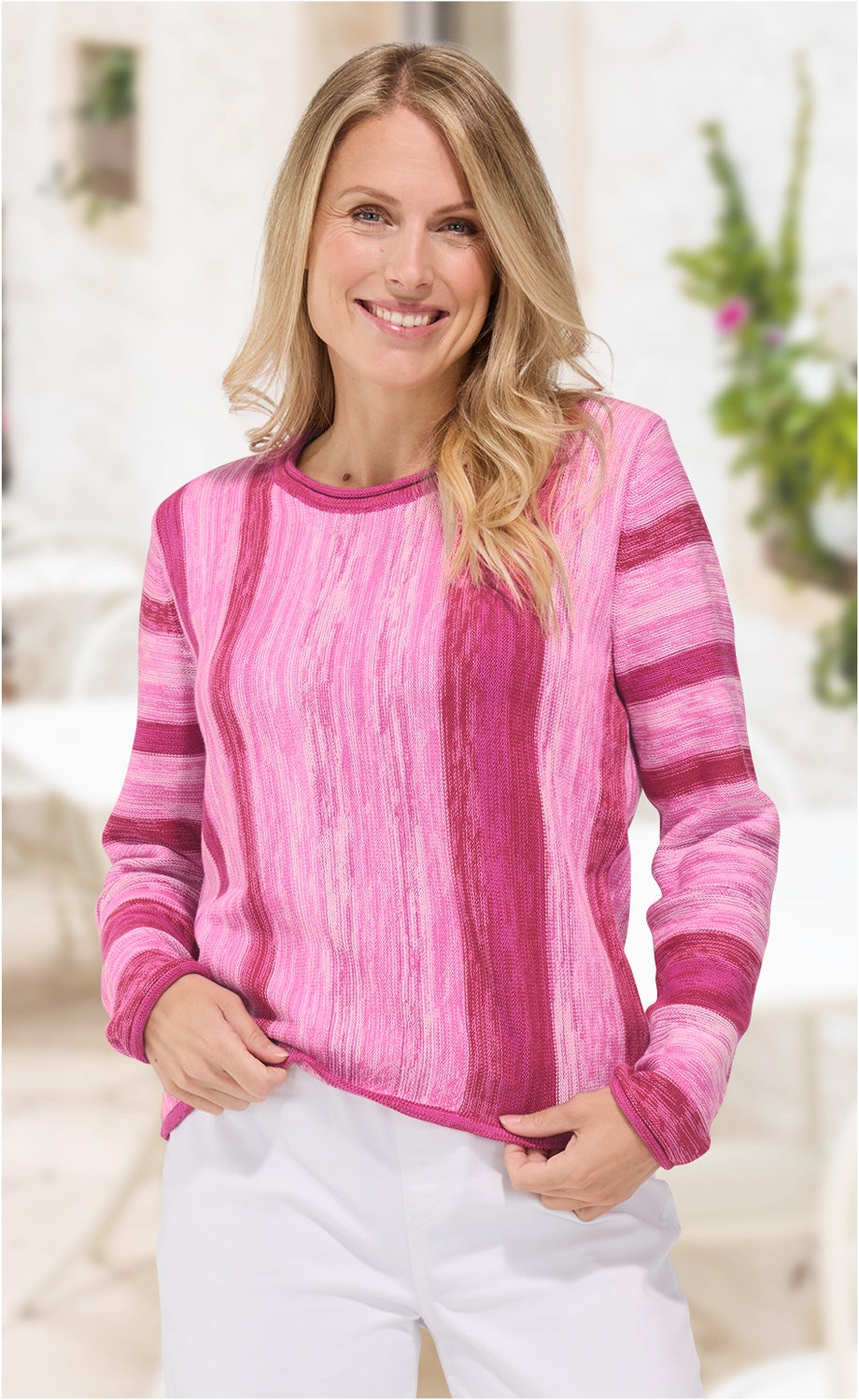 Frau mit langen blonden Haaren trägt pink-rosa gestreiften Baumwoll-Pullover mit Rundhalsausschnitt und weiße Hose, lächelt, hält Pullover leicht am Saum, unscharfer heller Hintergrund mit Grünpflanzen