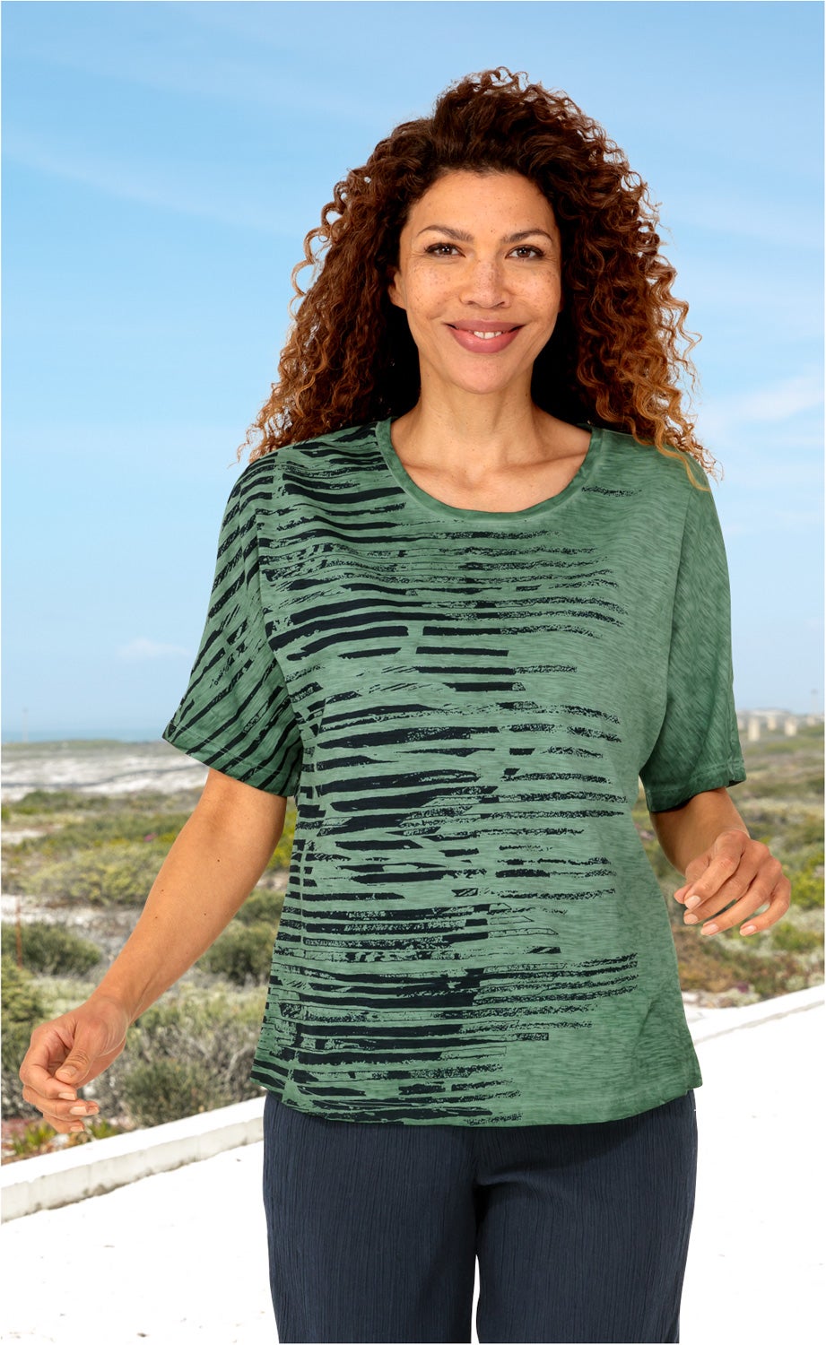 Frau mit lockigen Haaren trägt grünes Baumwoll-Shirt mit schwarzem Streifenmuster, steht draußen bei blauem Himmel und Vegetation, schwarze Hose, lächelnd