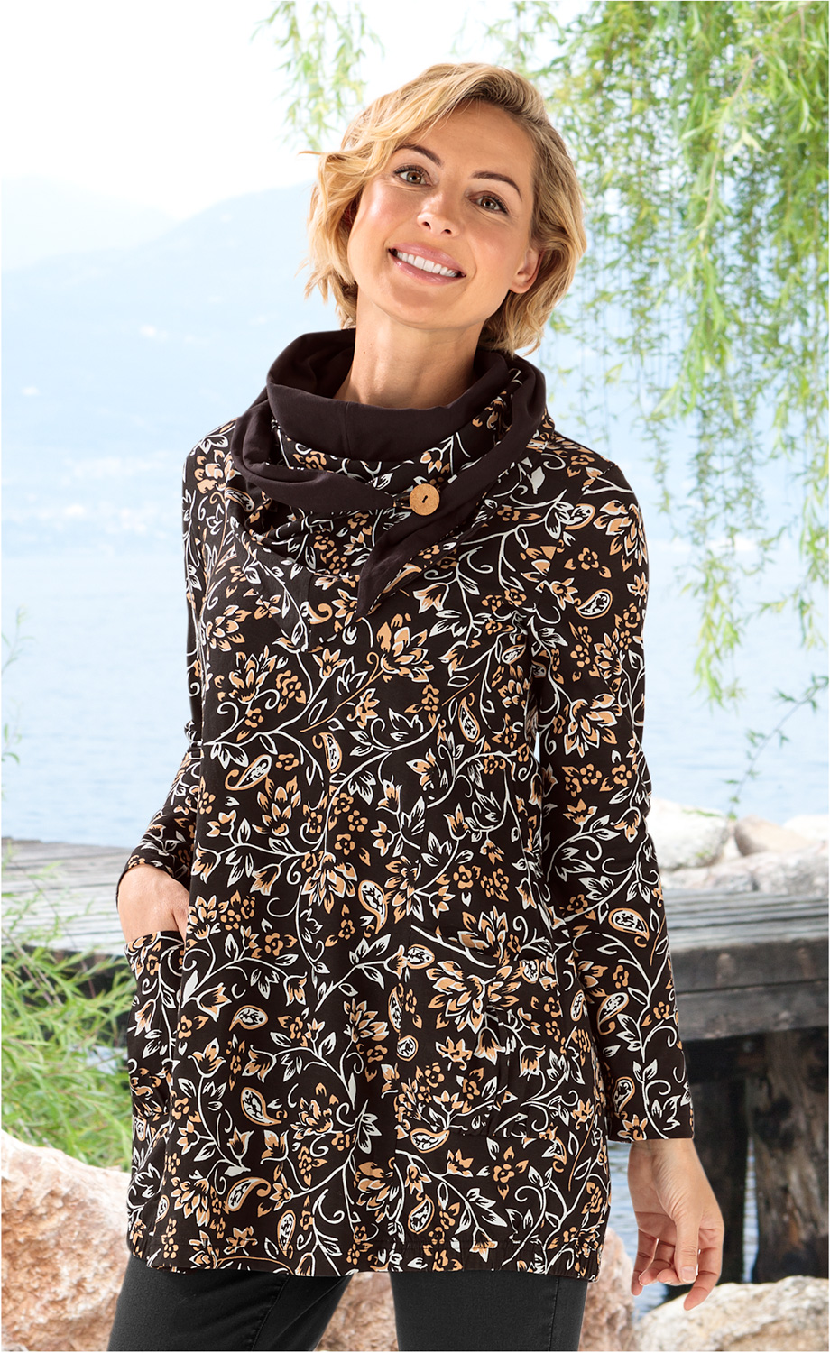 Frau trägt schwarzes Baumwoll-Shirt mit beige-weißem Blumenmuster und weitem Kragen mit Knopf, steht lächelnd vor See mit Bäumen und Steg im Hintergrund