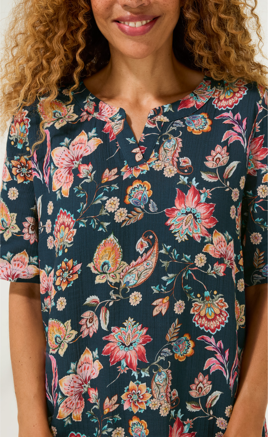 Baumwoll-Kleid mit dunklem Grund und buntem floralen Muster, V-Ausschnitt mit kleinem Schlitz, lockere kurze Ärmel, Nahaufnahme Oberkörper einer Frau mit lockigen Haaren und leichtem Lächeln.