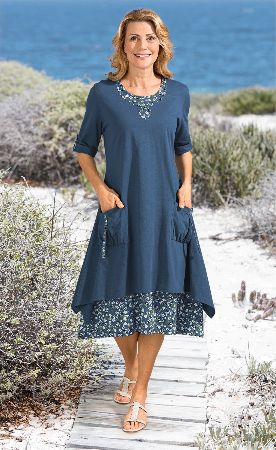 Frau in blauem Baumwoll-Kleid mit floralem Muster am Saum und Halsausschnitt, 3/4-Ärmeln, Taschen, steht auf Holzsteg am Strand vor Meer, trägt weiße Sandalen, lächelnd, sommerliche Atmosphäre