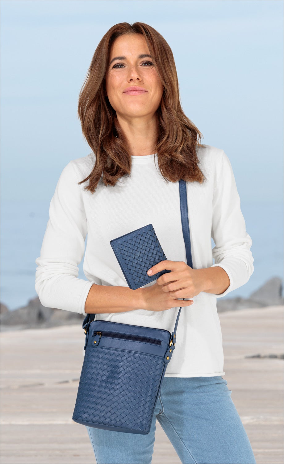 Frau mit weißen Pullover und blauen Jeans trägt eine blaue Umhängetasche 23x21x5cm mit geflochtenem Muster und hält ein passendes geflochtenes Portemonnaie in der Hand, Hintergrund Strand mit Felsen und blassem Himmel