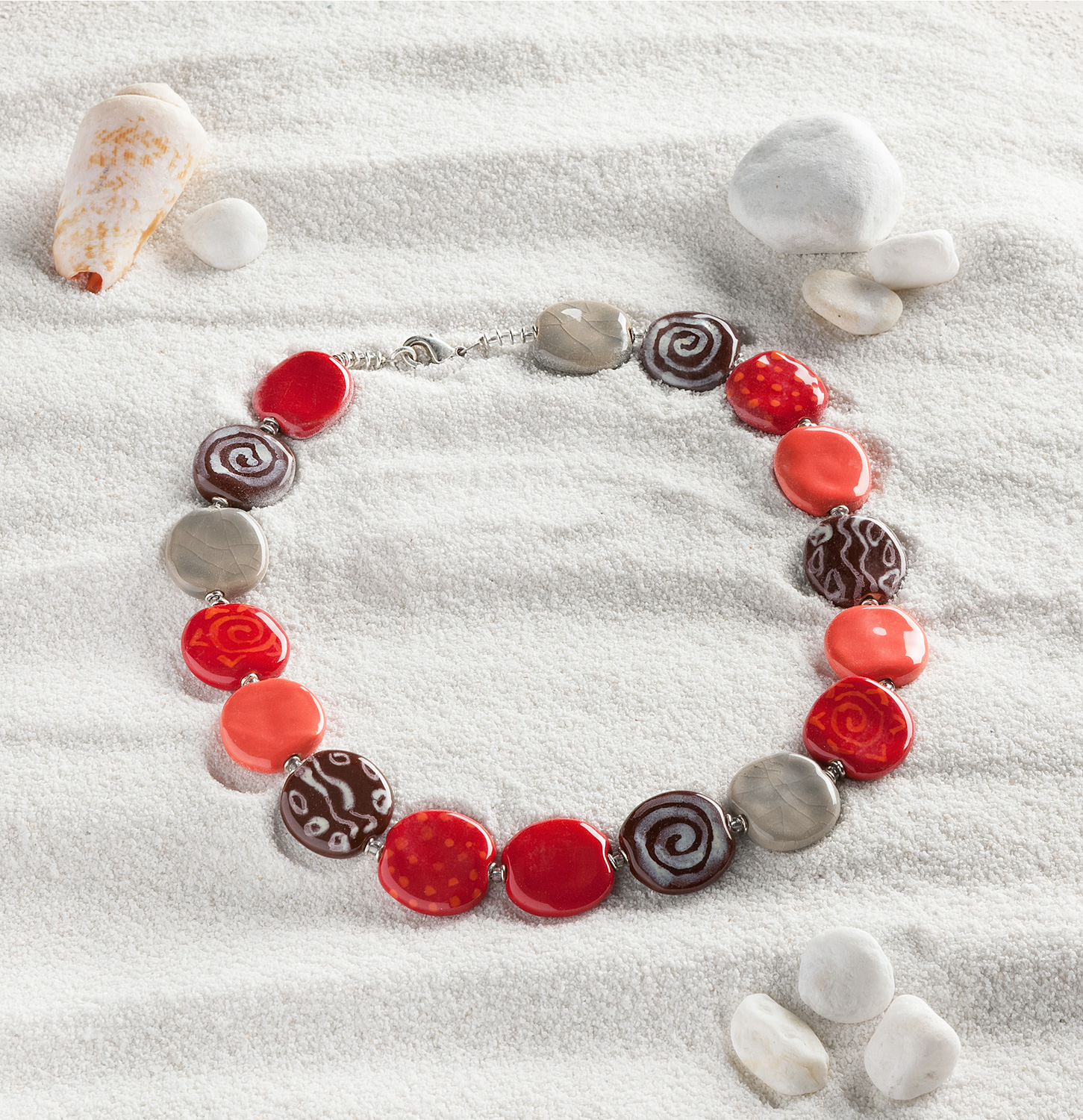 Keramik-Kette mit flachen, rundlichen Perlen in Rot, Grau und Braun mit verschiedenen Mustern, gelegt auf hellem Sand mit Muscheln und weißen Steinen als Dekoration.