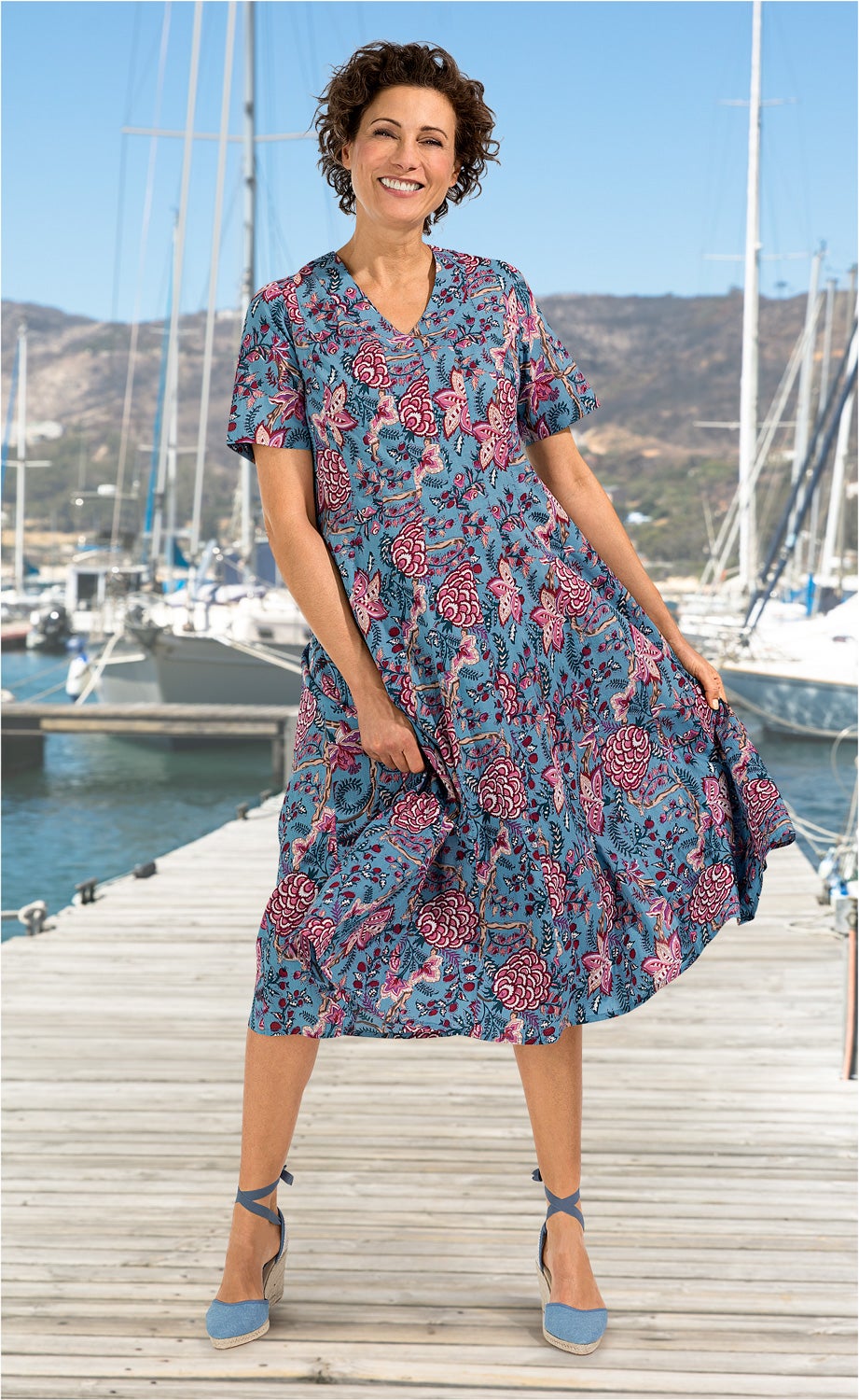 Frau trägt blaues Baumwoll-Kleid mit floralem Muster in Rot und Pink, steht lachend auf Holzsteg mit Segelbooten und Bergen im Hintergrund, trägt hellblaue Espadrilles mit Knöchelband