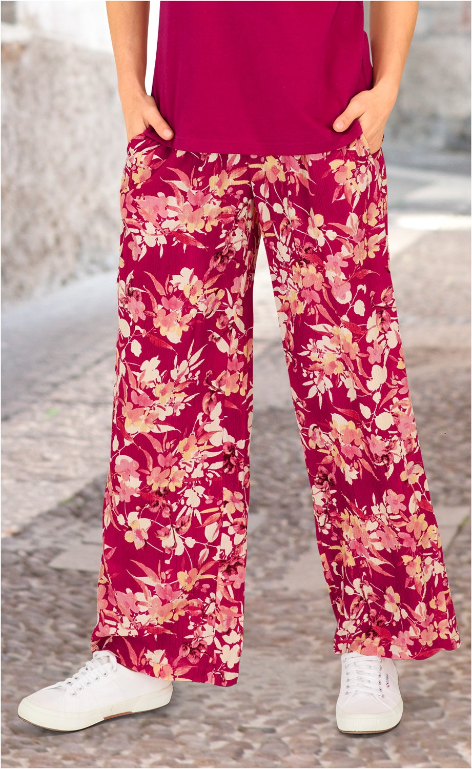 Beine in weiten Viskose-Hosen mit rosa und cremefarbenem Blumenmuster, kombiniert mit weißen Sneakern und einem dunkelroten Oberteil, stehend auf gepflastertem Boden