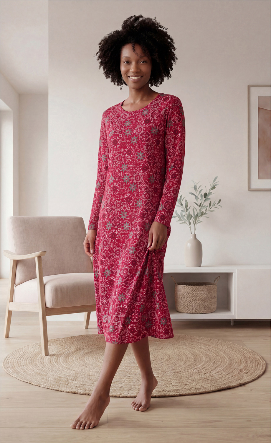 Frau mit lockigem Haar in rotem, langärmligem, knielangem Kleid mit floralem Muster, barfuß stehend in hellem Raum mit beige-grauem Sessel, rundem Naturfaserteppich, weißem Sideboard, Vase mit Pflanze und Wandbild