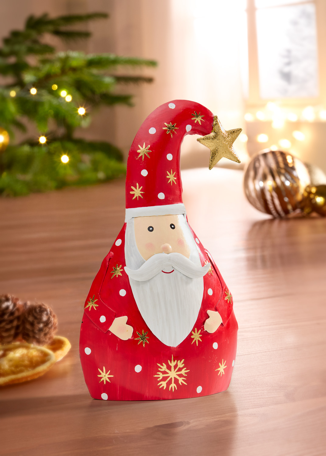 Roter Metall-Weihnachtsmann mit weißem Bart, roter Mütze mit goldenen Sternen und weißem Punktmuster, goldener Stern an der Mützenspitze, dekorativ auf Holztisch vor Weihnachtsbaum und Lichterkette