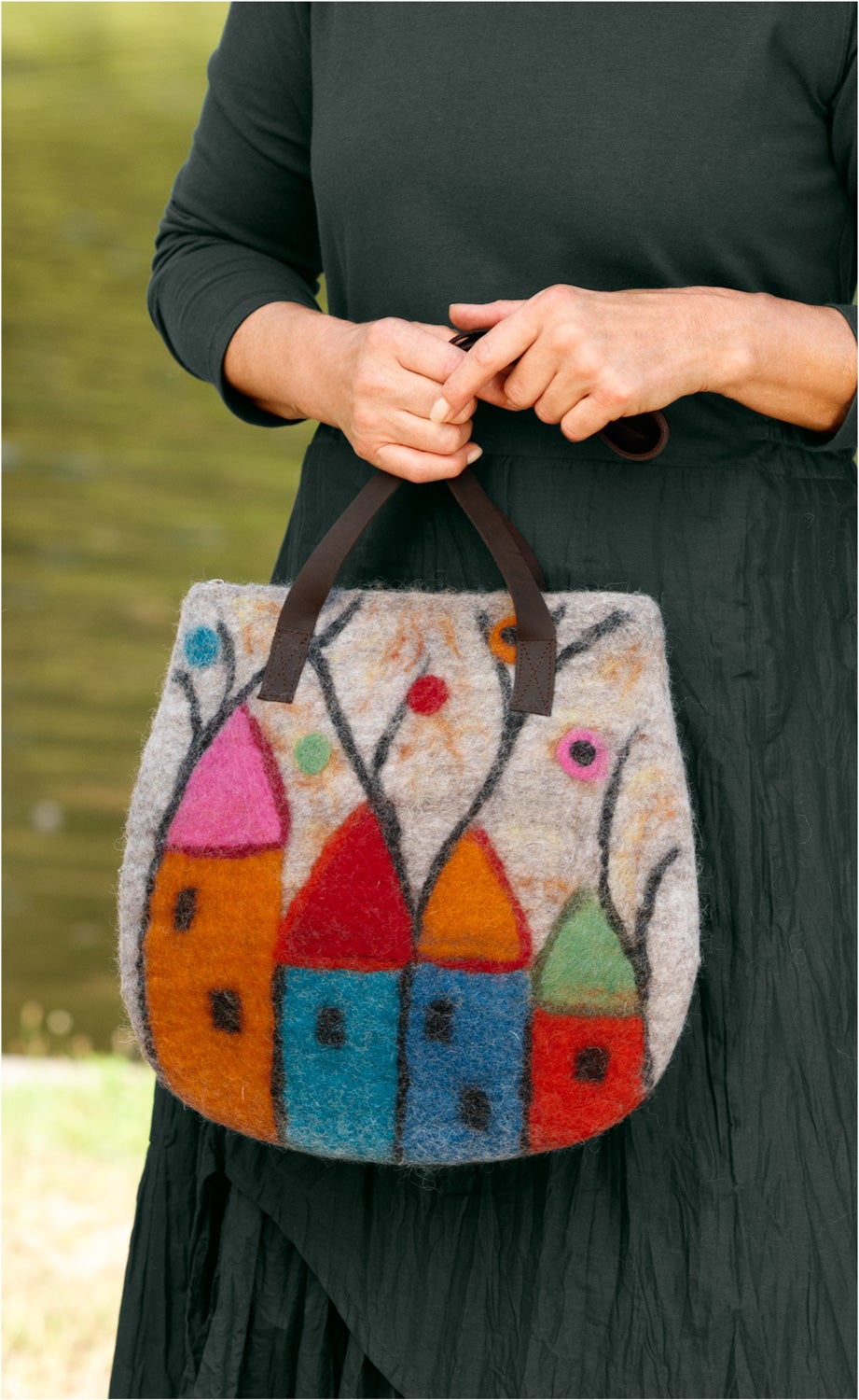 Frau in schwarzem Kleid hält bunte Filztasche mit Häuser- und Baum-Motiv, braune Lederriemen, unscharfer Hintergrund mit Wasser und Wiese