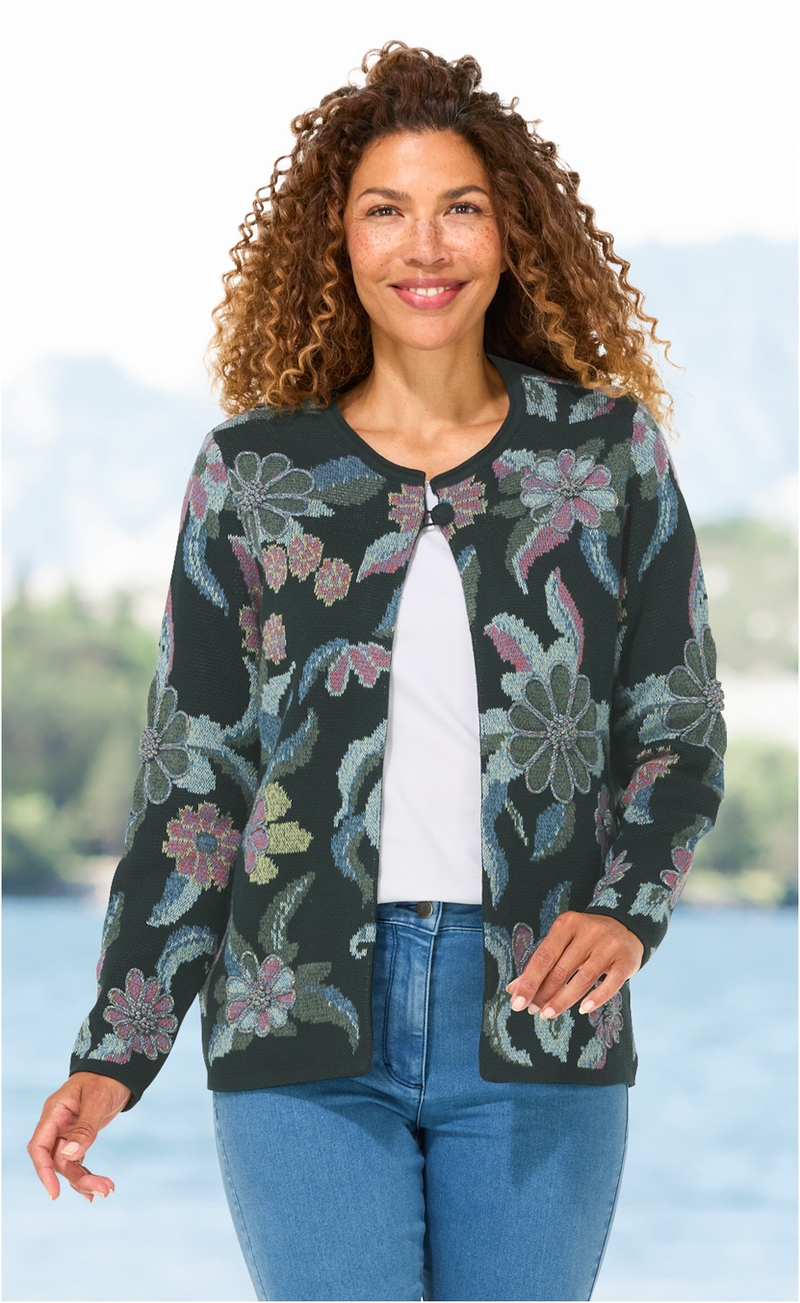 Frau trägt dunkelgrüne Baumwoll-Strickjacke mit buntem Blumenmuster, weißes T-Shirt und blaue Jeans, lächelnd, vor verschwommenem Fluss- und Berghintergrund