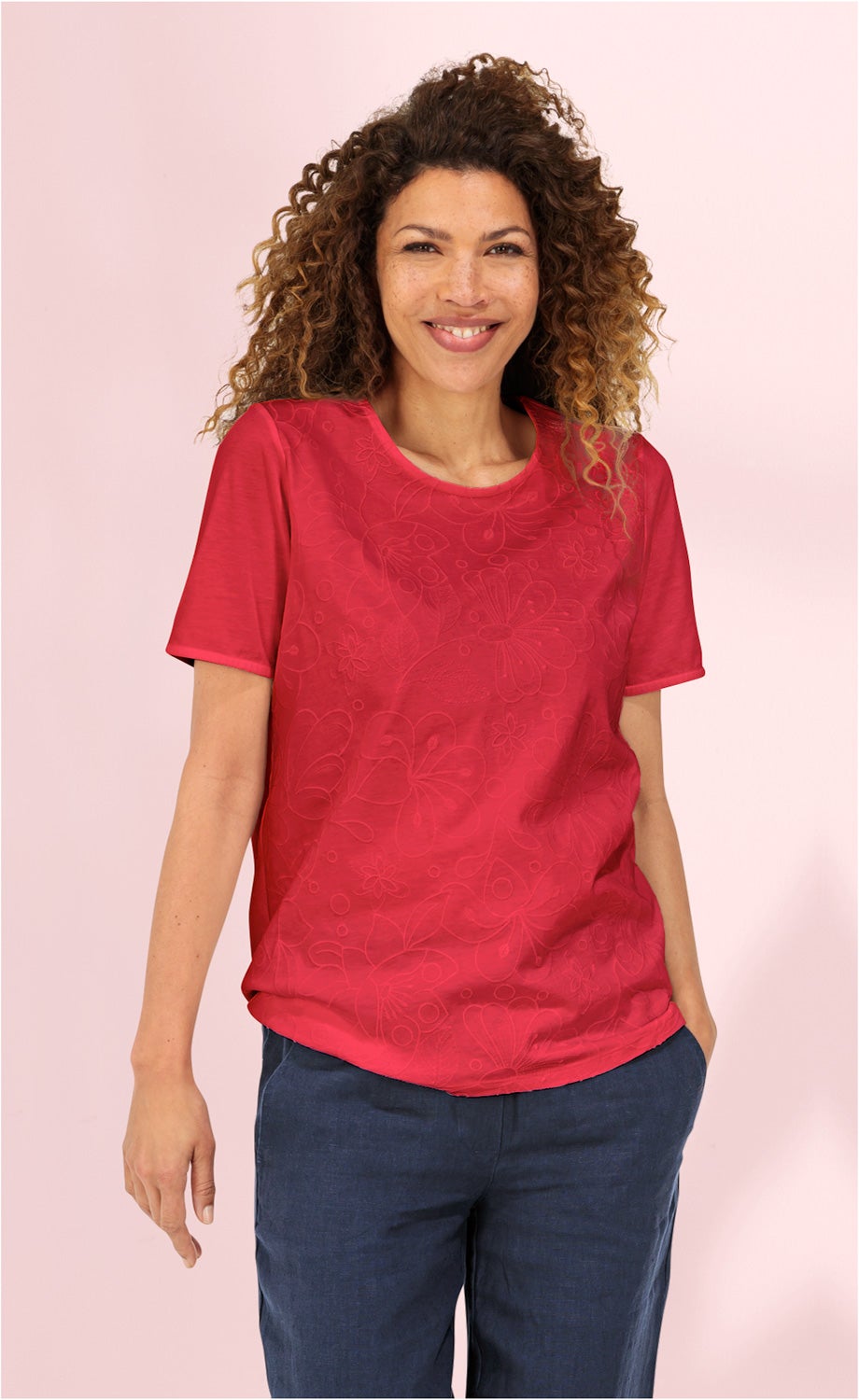 Frau mit lockigen Haaren trägt rotes Baumwoll-Shirt mit floralem Muster und blaue Hose, steht vor rosa Hintergrund und lächelt entspannt, eine Hand in der Hosentasche