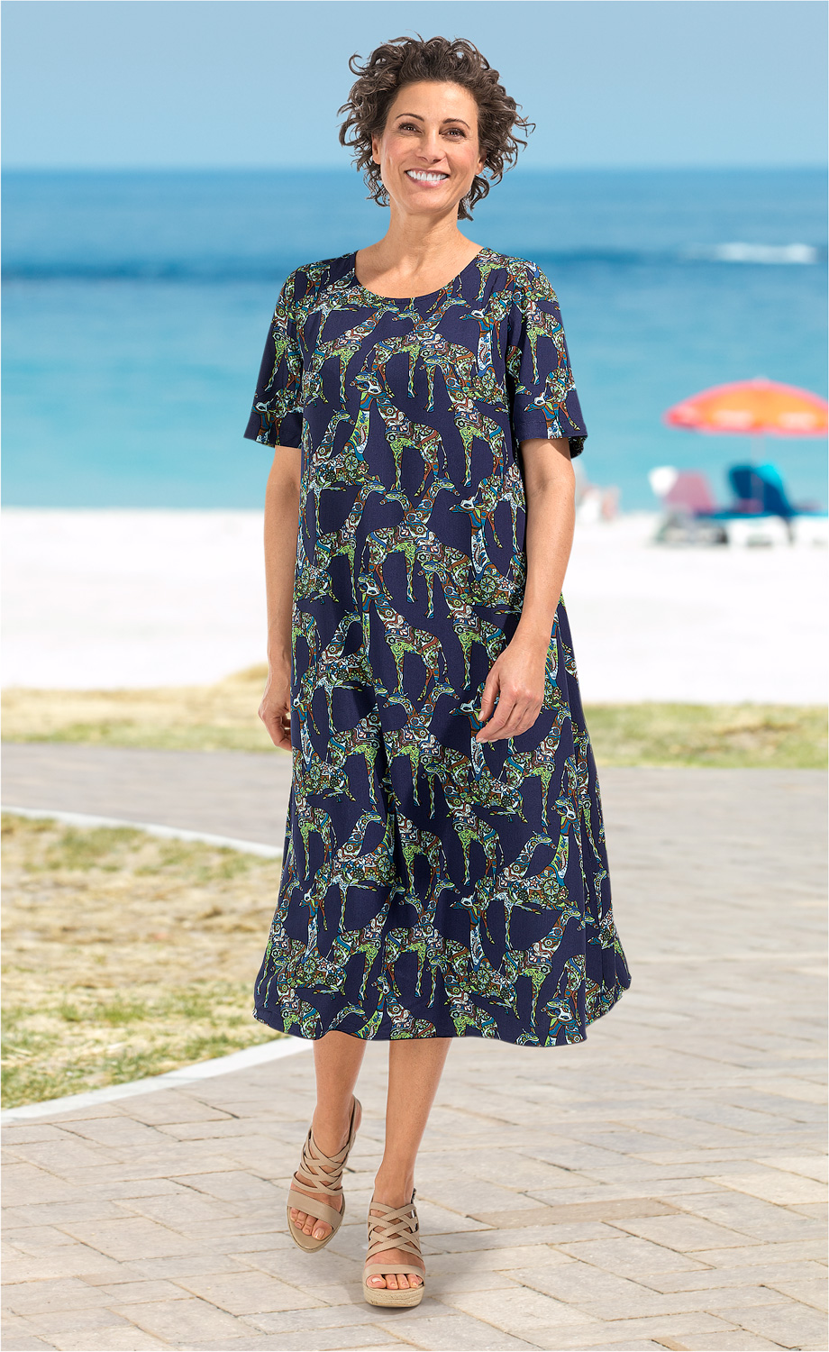 Frau trägt blaues Viskose-Kleid mit buntem Giraffen-Muster, kurze Ärmel, knöchellang, Sandalen, steht am Strandweg mit Meeresblick und buntem Sonnenschirm im Hintergrund, lächelt, kurze lockige Haare