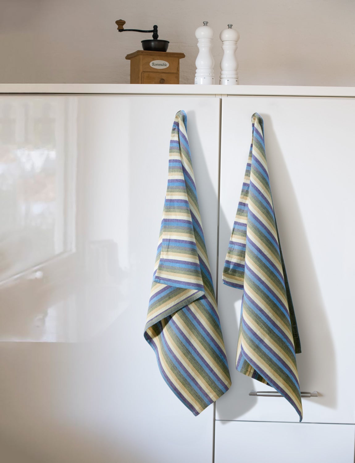 Zwei Baumwoll-Geschirrtücher mit diagonal verlaufenden Streifen in Blau, Grün, Beige und Weiß hängen an weißen Küchenschranktüren, darüber stehen eine alte Holz-Kaffeemühle und zwei weiße Gewürzmühlen.